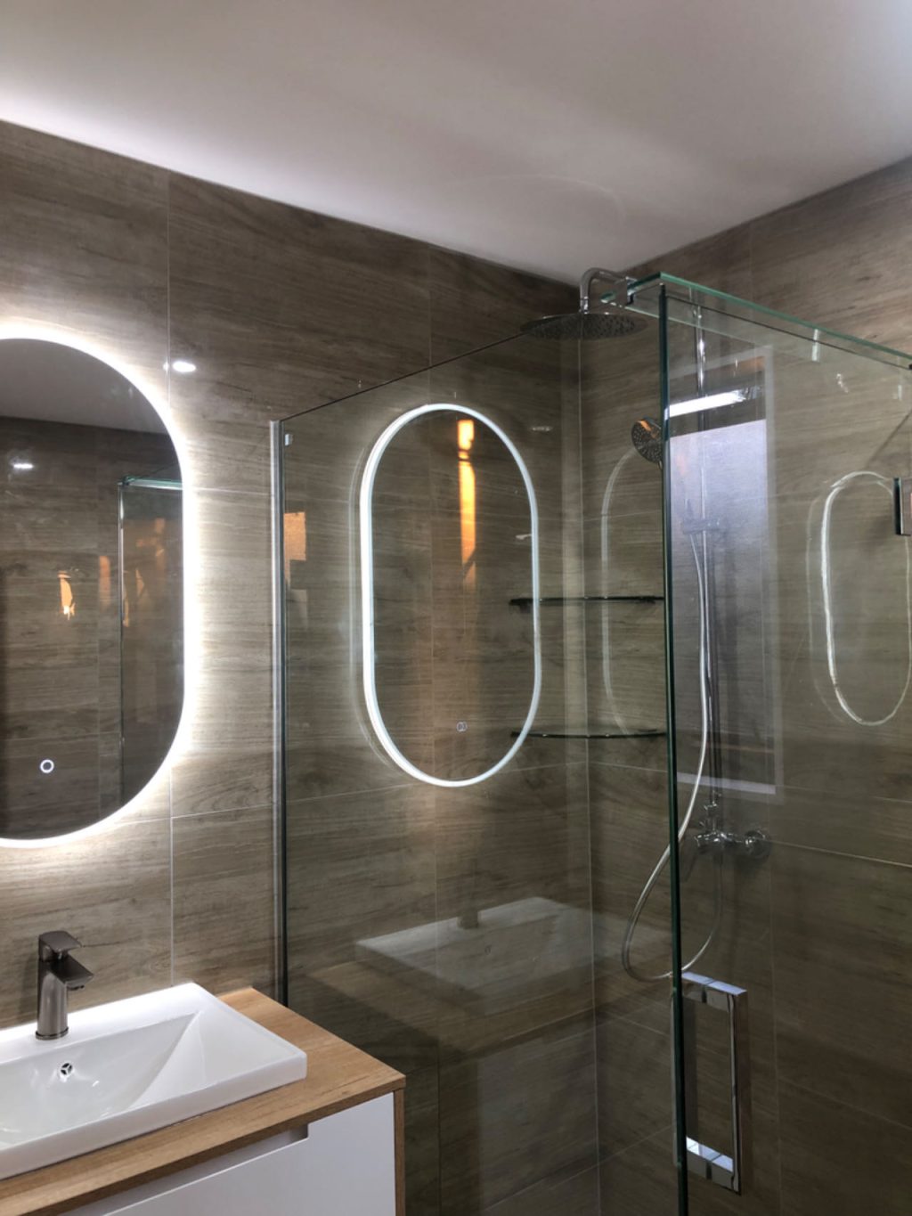 Modern bathroom with glass shower and illuminated oval mirror.