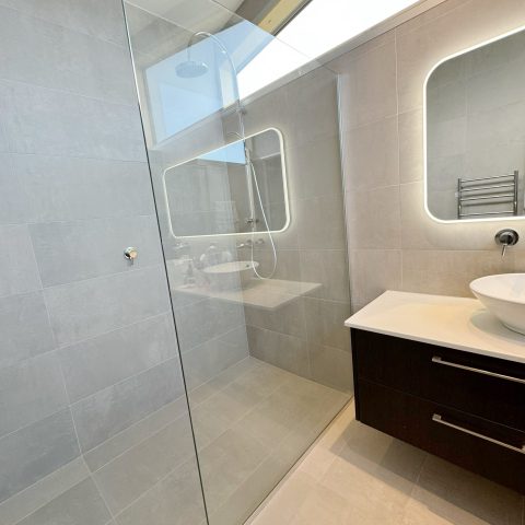 Modern bathroom with glass shower, illuminated mirror, and dark vanity with vessel sink.