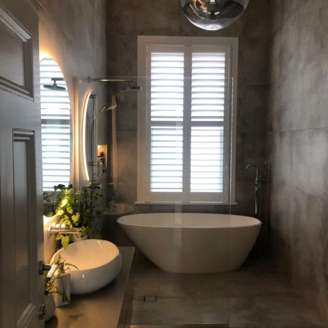 Modern bathroom with freestanding tub, round mirror, and window shutters.
