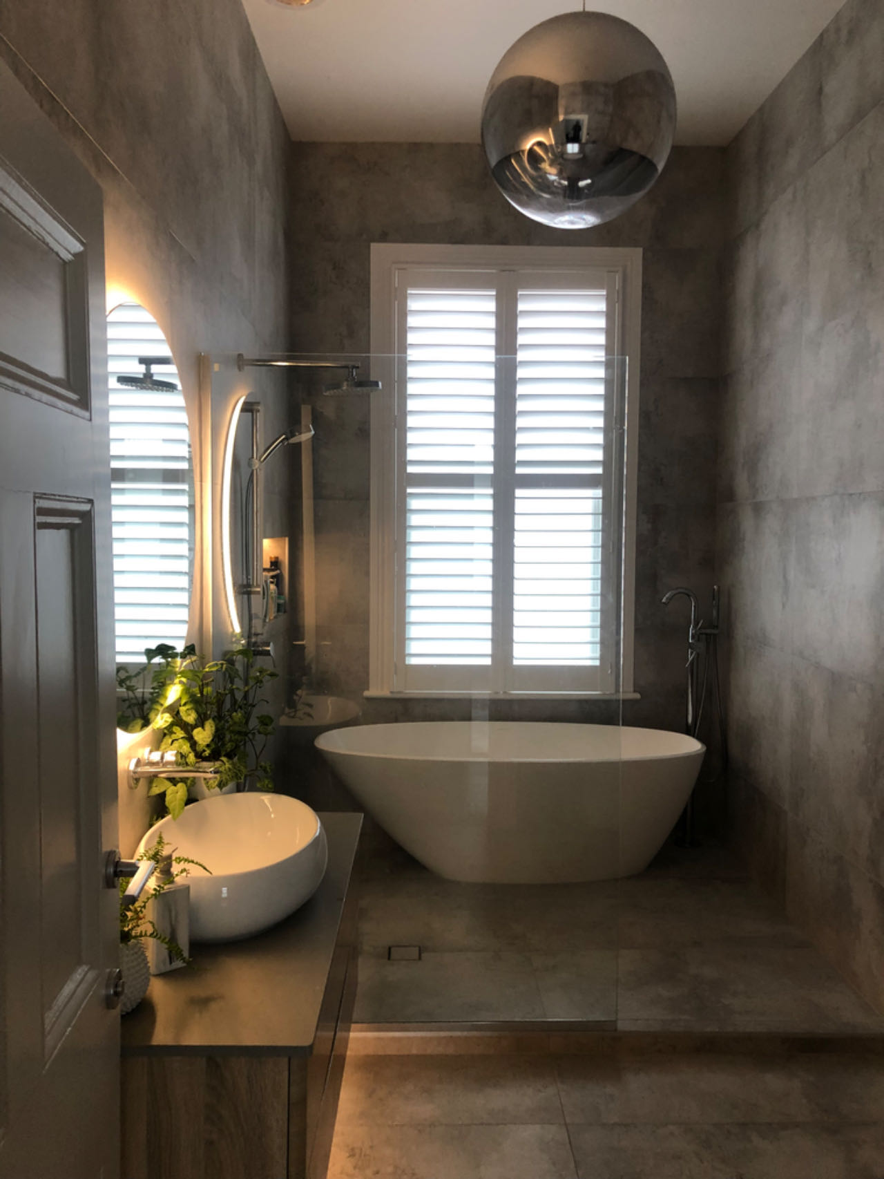 Modern bathroom with freestanding tub, round mirror, and window shutters.