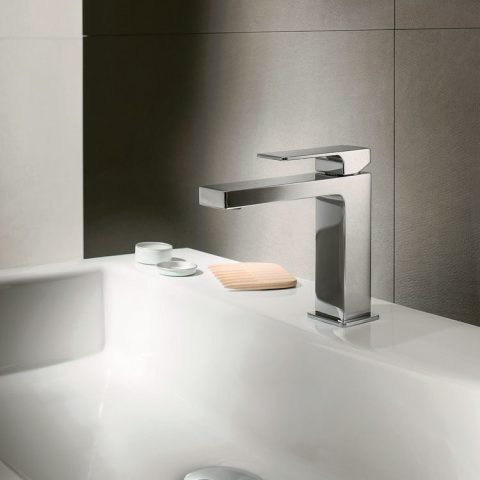Modern chrome faucet on white bathroom sink with soap and cups.