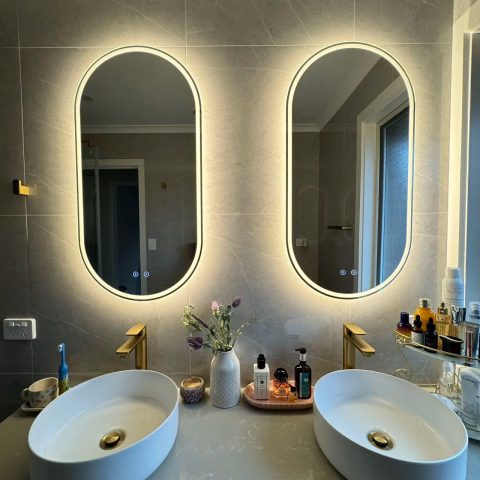 Modern bathroom with two oval mirrors, illuminated, above white sinks and gold faucets.