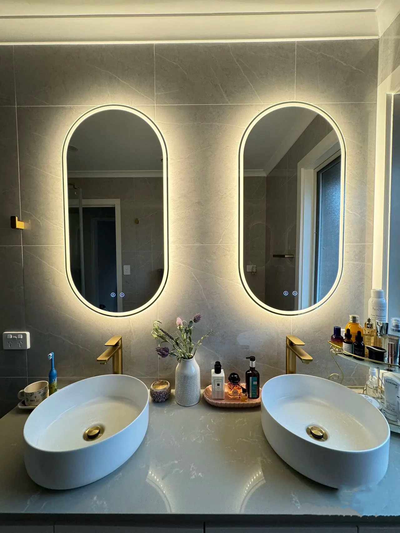Modern bathroom with two oval mirrors, illuminated, above white sinks and gold faucets.