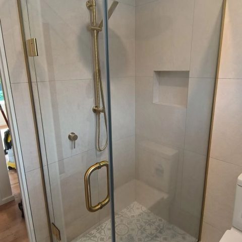 Modern shower with glass door, gold fixtures, and built-in shelf.